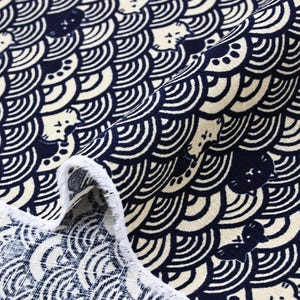 Puede incluir: Tela azul marino y crema con un patrón repetido de caras de gato estilizadas y diseños en forma de olas. La tela está doblada, revelando un borde blanco. Adecuada para diversos proyectos de costura y manualidades.