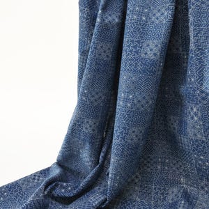 May include: A draped indigo fabric with a white geometric pattern. The fabric has a textured appearance and is folded, creating shadows and depth. The design features a repeating pattern of squares, stars, and zigzags, giving it a handcrafted look.