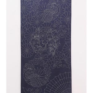 May include: A dark blue fabric wall hanging featuring white sashiko-style embroidery of two cranes, stylized floral patterns, a folding fan, and a group of seven figures. The textile is suspended from a light-colored wooden rod with a thin cord.