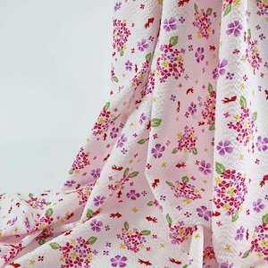 Puede incluir: Una tela blanca con una superficie texturizada y fruncida que presenta un patrón repetitivo de racimos de hortensias rosas y moradas, pequeñas hojas verdes, detalles amarillos y pequeños peces dorados rojos.