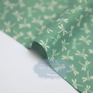 Puede incluir: Una tela de color verde salvia con un patrón repetitivo de pequeñas siluetas de libélulas blancas estilizadas. El material tiene una textura sutil de grano fino. Un logotipo azul claro con el texto DIY DISTRICT es visible en la tela.