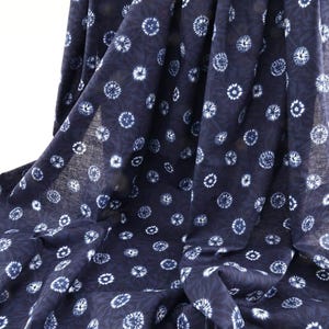 May include: Dark navy blue fabric with a repeating pattern of white, stylized floral or starburst designs. The fabric is draped, showing the texture and flow of the material. The pattern is evenly spaced across the fabric.