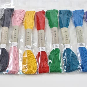 May include: A set of nine skeins of Sashiko embroidery thread in various colors including gray, black, pink, yellow, red, green, teal, blue, and purple. Each skein is individually wrapped in clear plastic and features a paper label with Japanese text.