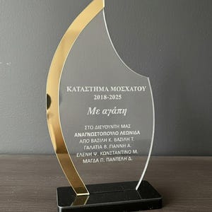 May include: A clear acrylic award with a gold-colored trim and a black base. The award features Greek text, including "ΚΑΤΑΣΤΗΜΑ ΜΟΣΧΑΤΟΥ 2018-2025" and names. It stands upright on a dark surface.