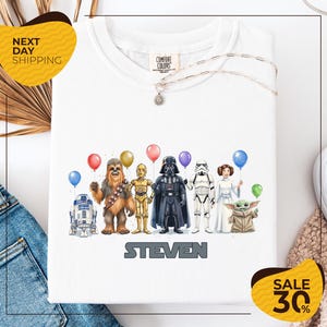 A white t-shirt with a graphic design featuring various characters from the Star Wars franchise, including Chewbacca, Darth Vader, Stormtrooper, and the character Steven. The characters are holding balloons, and the text "STEVEN" is displayed below the image.
