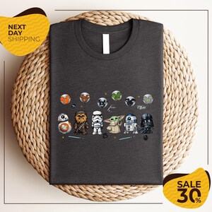 a gray t-shirt with a graphic design of various Star Wars characters, including Chewbacca, Stormtrooper, Darth Vader, and Baby Yoda, arranged in a row.