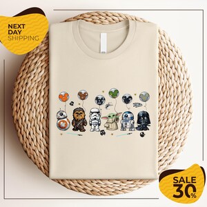 a beige t-shirt with a graphic design of various Star Wars characters, including Chewbacca, Stormtrooper, Darth Vader, and Baby Yoda, along with other Star Wars-themed elements.
