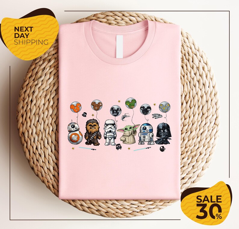A pink t-shirt with a graphic design featuring various Star Wars characters, including Chewbacca, Stormtrooper, Darth Vader, and Baby Yoda, along with other Star Wars-themed elements.