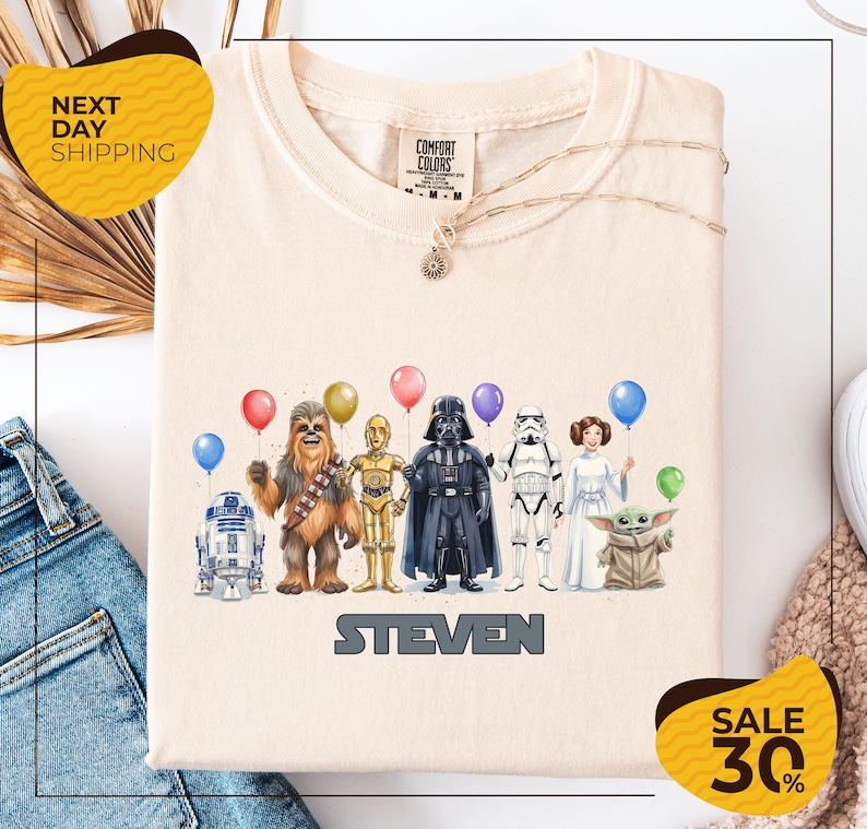a t-shirt with a graphic design of various Star Wars characters, including Chewbacca, Darth Vader, Stormtrooper, and Yoda, along with balloons.