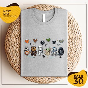 A gray t-shirt with a graphic design featuring various Star Wars characters, including Chewbacca, Stormtrooper, Darth Vader, and Baby Yoda, along with balloons.