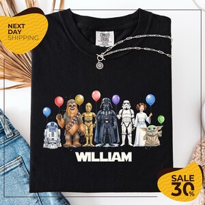 A black t-shirt with a graphic design featuring characters from the Star Wars franchise, including Chewbacca, Darth Vader, Stormtrooper, and Yoda, along with colorful balloons.
