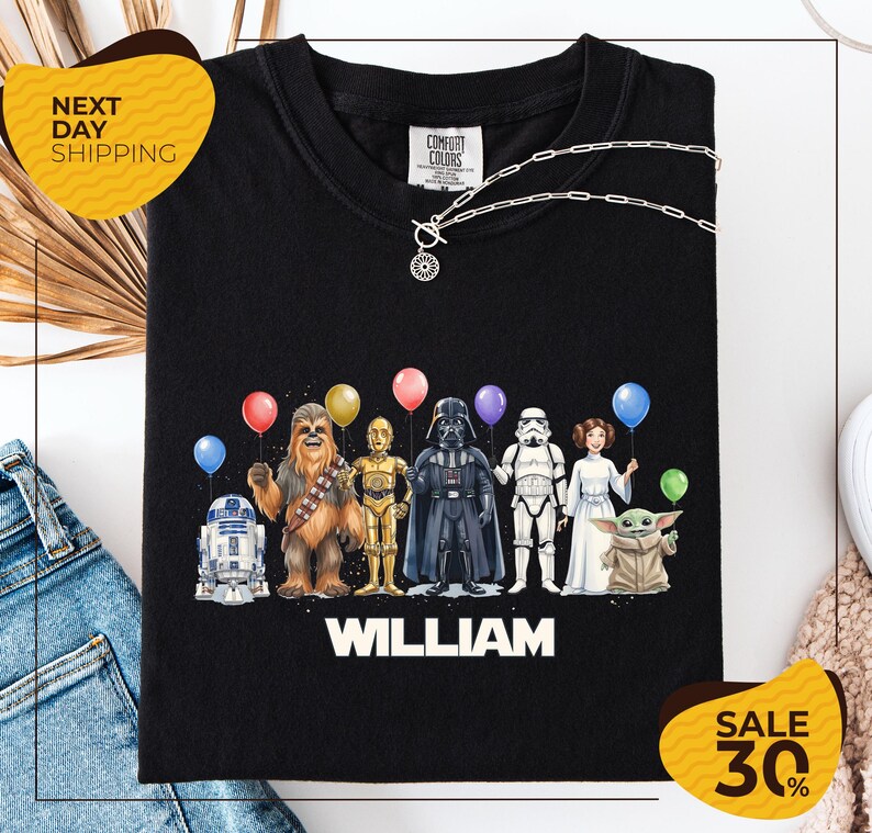 A black t-shirt with a graphic design featuring characters from the Star Wars franchise, including Chewbacca, Darth Vader, Stormtrooper, and Yoda, along with colorful balloons.