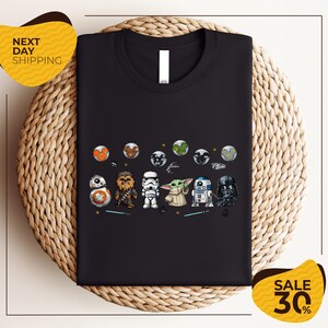 a black t-shirt with a graphic design of various Star Wars characters, including the main characters from the Star Wars franchise, such as Rey, Finn, and Chewbacca, along with other iconic characters like Bond, Baby Yoda, and Darth Vader. The t-shirt is displayed on a woven placemat, and there are two yellow sale signs in the image, indicating a discount or sale event.