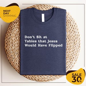 May include: Navy blue t-shirt with the text "Don't Sit at Tables that Jesus Would Have Flipped" in white. The shirt is displayed on a woven, light brown surface. A yellow sticker with "Next Day Shipping" is in the upper left corner.