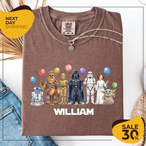 A brown t-shirt with a graphic design featuring characters from the Star Wars franchise, including Chewbacca, Darth Vader, Stormtrooper, and Yoda, along with colorful balloons. The text "WILLIAM" is displayed below the characters, and there are two yellow sale signs indicating a 30% discount on the shirt.