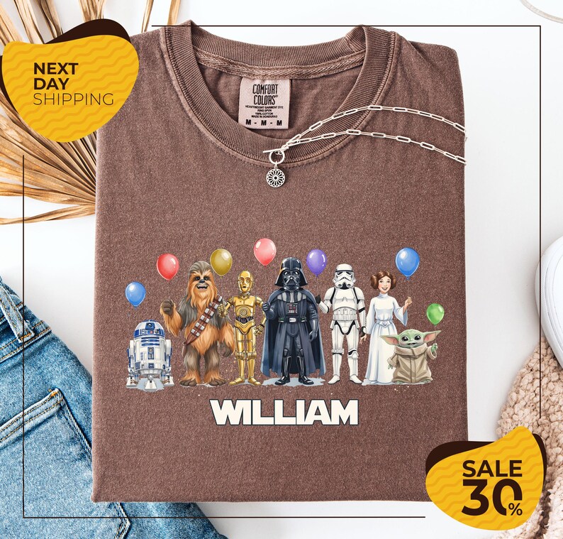 A brown t-shirt with a graphic design featuring characters from the Star Wars franchise, including Chewbacca, Darth Vader, Stormtrooper, and Yoda, along with colorful balloons. The text "WILLIAM" is displayed below the characters, and there are two yellow sale signs indicating a 30% discount on the shirt.