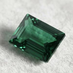Puede incluir: Una piedra preciosa rectangular de corte esmeralda con un color verde bosque profundo y superficies facetadas que reflejan la luz. La piedra presenta bordes afilados y limpios con un acabado transparente y pulido, mostrando una estructura interna clara.