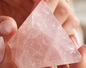 May include: A pale pink rose quartz crystal carved into a geometric pyramid shape. The stone features a translucent appearance with natural internal fractures and inclusions visible throughout its faceted surface, reflecting light across its smooth edges.