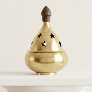 May include: A gold-toned metal incense burner with a rounded base and a conical lid featuring star-shaped cutouts. The lid is topped with a small, dark brown wooden finial. The object has a smooth, polished finish and a horizontal line detail.