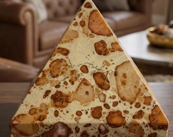 May include: A triangular pyramid sculpture crafted from polished stone featuring a speckled pattern of tan, beige, and dark brown circular and irregular inclusions embedded within a lighter cream-colored matrix, displaying a smooth, glossy finish.