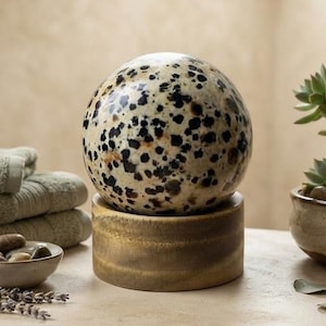 Puede incluir: Una piedra esférica con base color crema y manchas negras sobre un soporte cilíndrico de madera, junto a una pila de toallas dobladas de color verde salvia, un cuenco pequeño con piedras lisas y una planta suculenta en maceta.