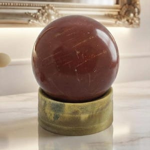 May include: A polished, deep red spherical stone orb resting on a cylindrical, weathered yellow-green base. The smooth, dark red sphere features subtle natural veining and sits centered on a textured, matte-finish stand with visible color variations.