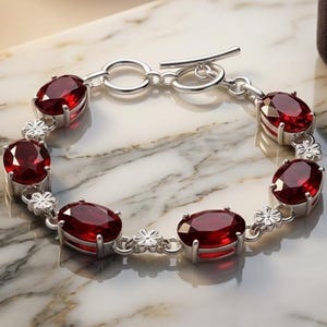 Könnte beinhalten: Ein silbernes Armband mit fünf oval geschliffenen, tiefroten Granat-Edelsteinen in Metallfassungen. Die Verbindungsglieder zwischen den Steinen sind als kleine, detaillierte Silberblumen gestaltet. Verschluss mit Knebel und Ring.