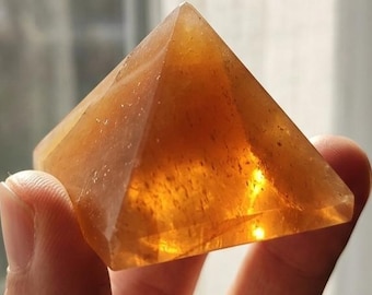 May include: A translucent, golden-orange pyramid-shaped stone with a smooth, polished surface. The object exhibits internal natural inclusions and variations in color density, catching the light to create a warm, glowing effect throughout.