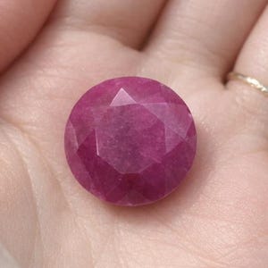 May include: A faceted, round, deep pink gemstone. The gem is held in a hand, showcasing its polished surface and geometric cuts. The stone's color is a vibrant, saturated pink, reflecting light across its many facets.
