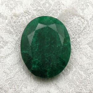 May include: A faceted, oval-shaped emerald gemstone. The gem is a deep, rich green color and is set against a white, patterned fabric. The cut of the stone creates multiple facets that reflect light.