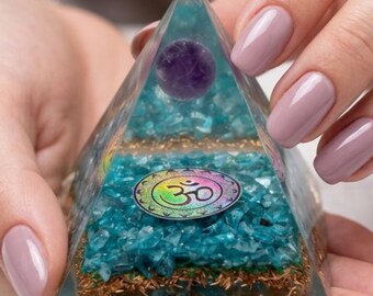 May include: A clear resin pyramid containing layers of blue crystal chips, copper shavings, and a single purple sphere at the top. A circular metallic emblem featuring the Om symbol in a rainbow gradient is embedded on the front face of the pyramid.