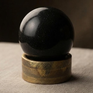 Side perspective of a smooth black tourmaline polished sphere supported by a golden-hued vintage metal ring base. High energy chakra healing stone ball suitable for witchy room aesthetic and feng shui altar decor.