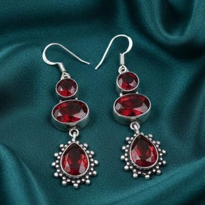 Puede incluir: Un par de pendientes colgantes plateados con tres piedras rojas cada uno. La piedra superior es redonda, la central ovalada y la inferior en forma de lágrima, rodeadas por un borde plateado decorativo. Los pendientes cuelgan de un gancho plateado.