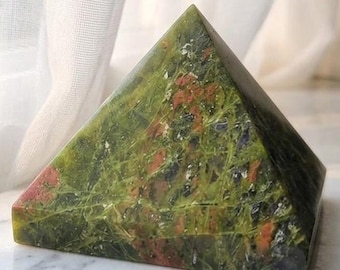 May include: A pyramid-shaped stone sculpture featuring a mottled surface with deep forest green, olive, and reddish-brown mineral inclusions. The object has a smooth, polished finish and displays a complex, natural pattern of varied colors throughout.