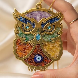 May include: A gold-toned owl pendant featuring a mosaic of colorful crushed stones including purple, blue, green, orange, and yellow segments. The owl has two bright blue eyes and a central evil eye bead at the bottom, suspended from a gold chain.