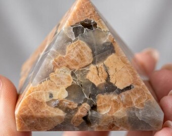 May include: A pyramid-shaped stone object featuring a polished surface with a mottled pattern of tan, beige, and translucent gray inclusions. The stone displays a network of fine, dark, and light-colored fractures throughout its geometric structure.