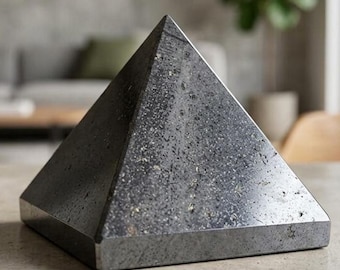 May include: A dark gray stone pyramid sculpture with a speckled, textured surface. The object features a smooth, polished finish with visible natural mineral inclusions throughout the material, forming a sharp geometric point at the top.