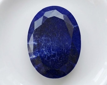 Puede incluir: Una gema ovalada, facetada, de color azul oscuro con vetas blancas, sobre un plato blanco. La superficie de la gema refleja la luz, mostrando su talla. La composición es sencilla, destacando el color y la forma de la gema.