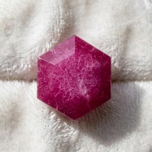 Könnte beinhalten: Ein facettierter, sechseckiger Edelstein in tiefem Magenta. Der Stein hat ein komplexes Innenmuster und liegt auf einer weichen, weißen, strukturierten Oberfläche. Form und Farbe des Steins deuten darauf hin, dass er ein dekoratives Element oder ein Schmuckstück sein könnte.