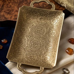 Op de afbeelding: Een rechthoekig koperen dienblad met een ingewikkeld reliëf bloemenpatroon over het oppervlak. Het dienblad heeft twee decoratieve handgrepen aan de uiteinden, een metallic gouden afwerking en een gedetailleerd gegraveerd ontwerp van bloemen.