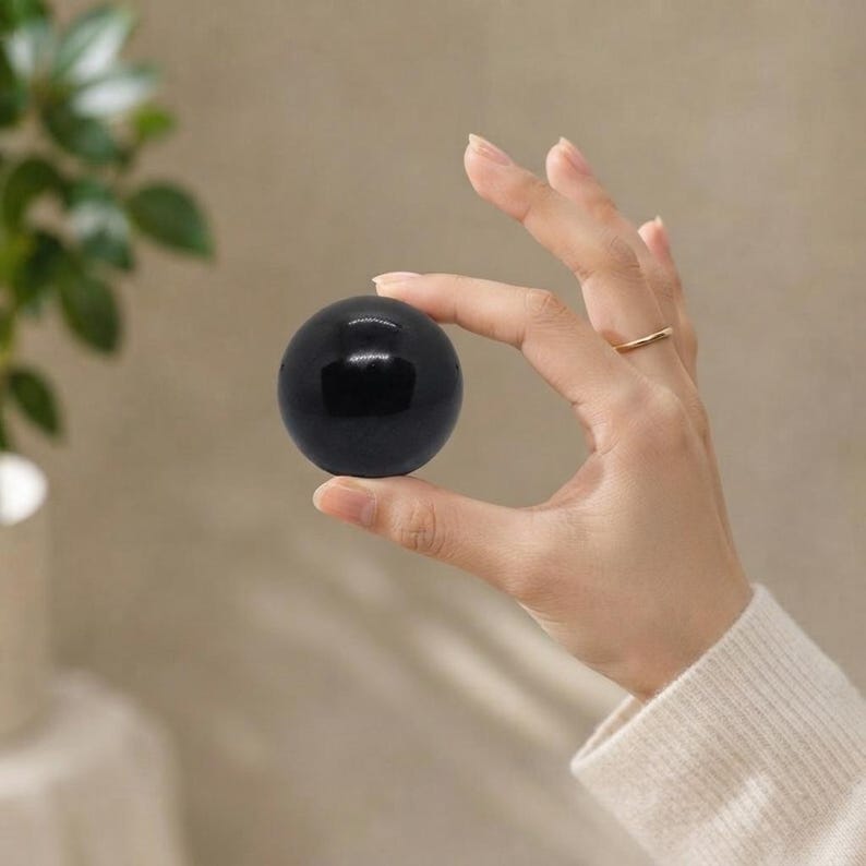 Detailed view of a hand-polished black tourmaline crystal ball with a metallic ring holder, placed on a warm neutral linen background. Excellent grounding gemstone sphere for emf protection and root chakra balancing.