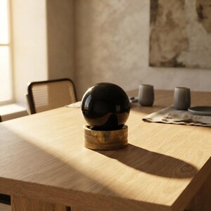 Deep black tourmaline crystal sphere on a brass display stand resting on a soft fabric mat under warm moody lighting. Beautiful gemstone orb offering electromagnetic shielding and spiritual grounding for desk workspace.