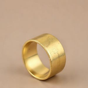Op de afbeelding: Een brede, goudkleurige ring met een licht getextureerd oppervlak. De ring heeft een eenvoudig, bandvormig ontwerp en staat tegen een neutrale achtergrond. De ring lijkt van metaal te zijn.