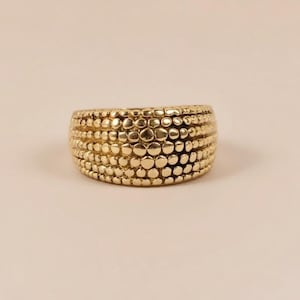 May include: A gold-colored ring with a textured surface. The ring is wide and features a pattern of small, raised circles across its surface. The ring is a warm gold color and appears to be made of metal.