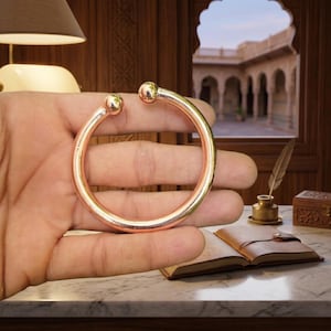 May include: A copper-toned bangle bracelet with rounded ball ends. The bracelet is held in a hand, showcasing its circular design. The jewelry is a simple, elegant accessory.