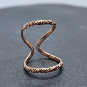 May include: A bronze-colored double-finger ring. The ring features an open design with two bands connected by a curved central element. Both bands are adorned with a repeating geometric pattern, adding texture and detail to the jewelry.