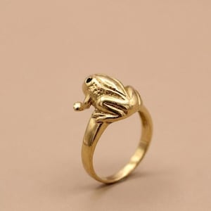 Könnte beinhalten: Ein goldfarbener Ring mit einem detaillierten Froschdesign. Der Frosch sitzt auf dem Ringband, mit strukturierter Haut und einem kleinen, runden Auge. Der Ring besteht aus einem glänzenden, metallischen Material.
