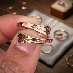 Puede incluir: Dos anillos de cobre martillado se sostienen cerca de la cámara. Los anillos tienen una superficie texturizada y un color marrón rojizo cálido. Otros anillos son visibles en el fondo, junto con una caja de anillos.