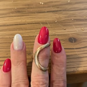 May include: A gold-coloured ring encircles a finger with a red nail. The ring has a curved, organic form. Other fingers display red and white nail polish. The background is a wooden surface.