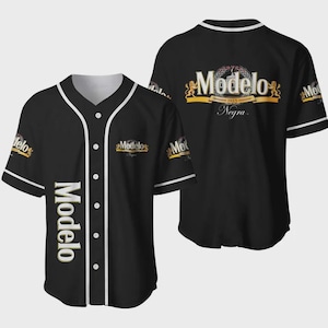May include: Black baseball jersey with white trim and buttons. The jersey features the Modelo logo in gold and white on the front and back. The back of the jersey also displays the words "Modelo Negra" and the year "1925".